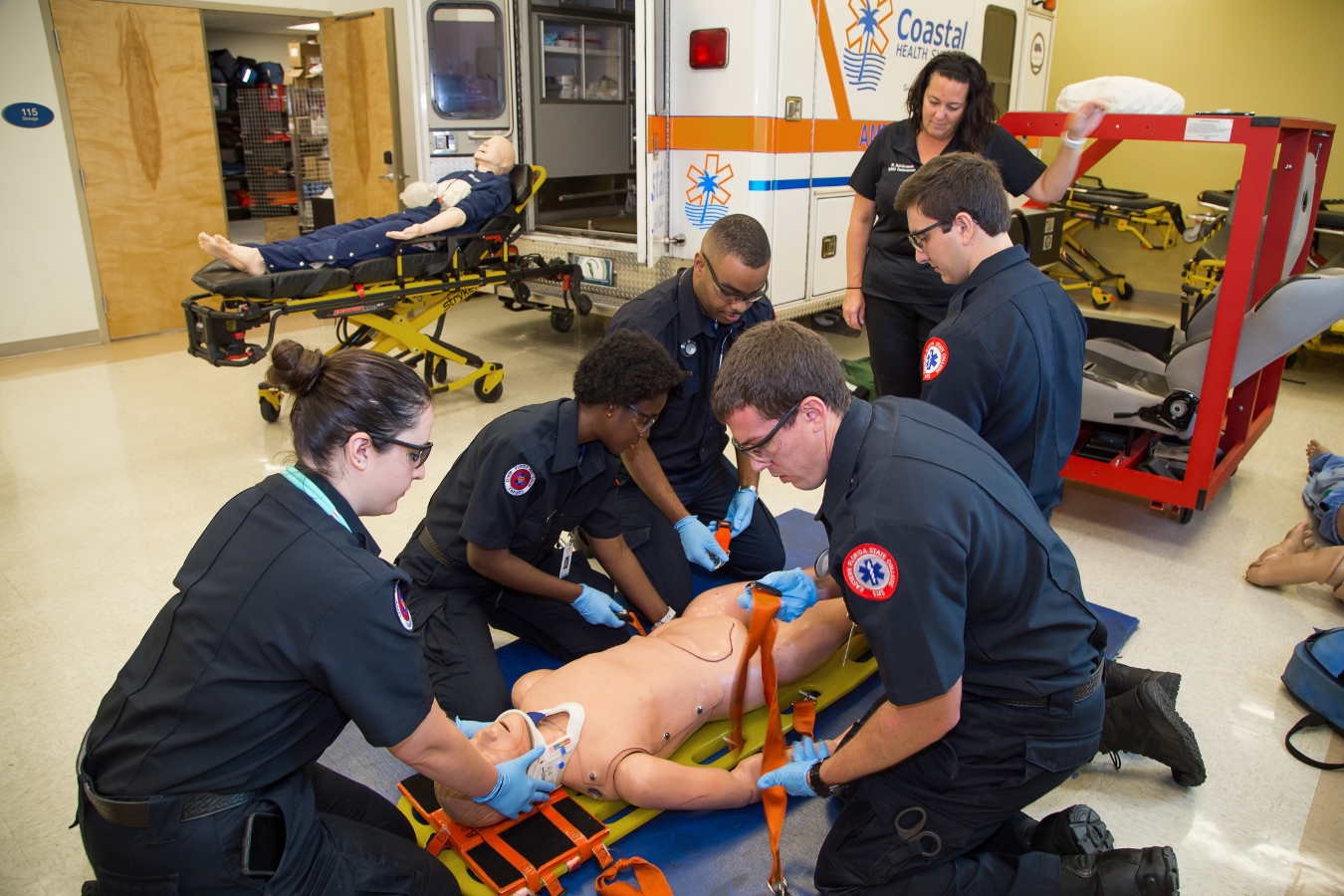 Diverse group of EMS/EMT students training in an environment with an ambulance, human patient simulation dummies, real-world tools, and instructor.