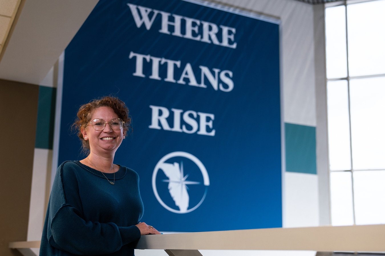 Woman with red hair smiling in front of a blue banner that says, "Where Titans Rise".