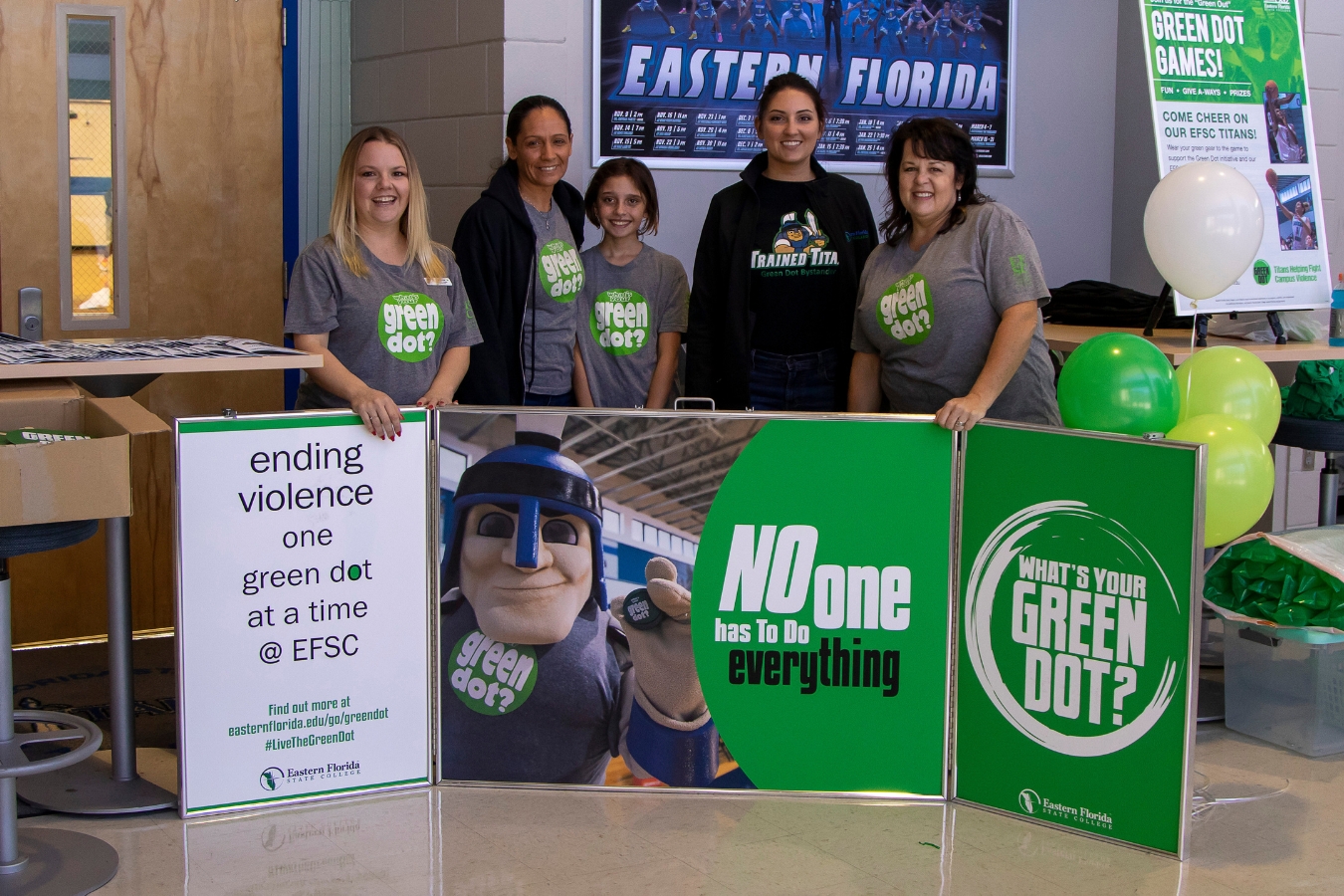 Green Dot presenters and community representatives pose for a photo holding a Green Dot Banner that says, "No one has to do everything", "What's your Green Dot?", and "Ending violence, one green dot at a time".