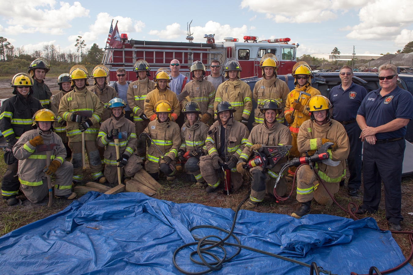 Group photo of firefighter training recruit group posing for a photo with their instructors, tools, and gear in front of a fire truck. 