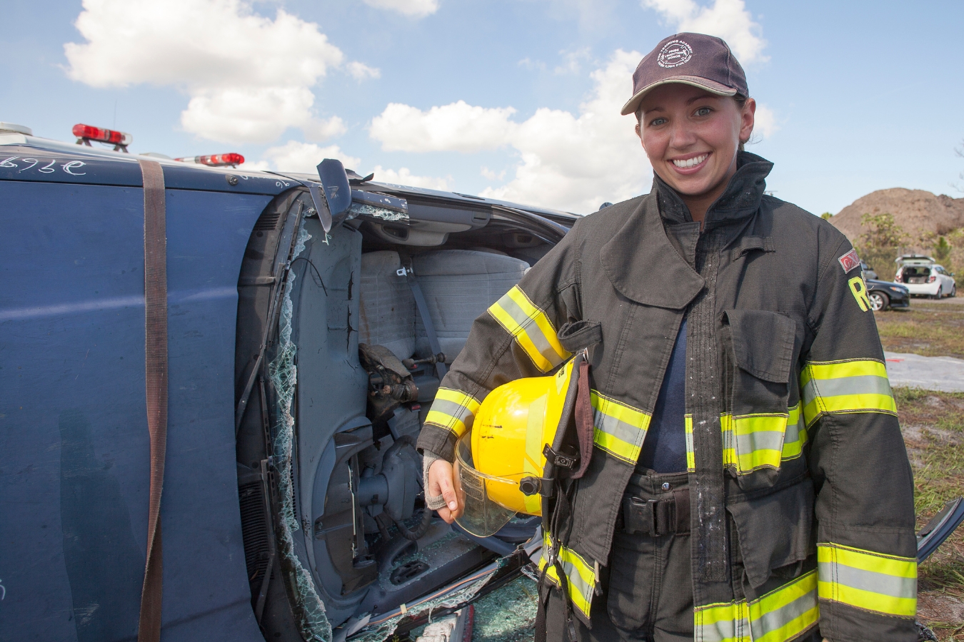 firefighter student poses in front of a car without its room with a smile while holding her helmet