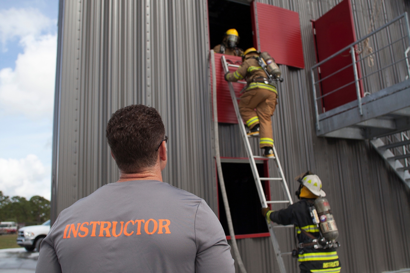 An instructor looks on as students work together to climb a ladder into a window of the fire academy tower