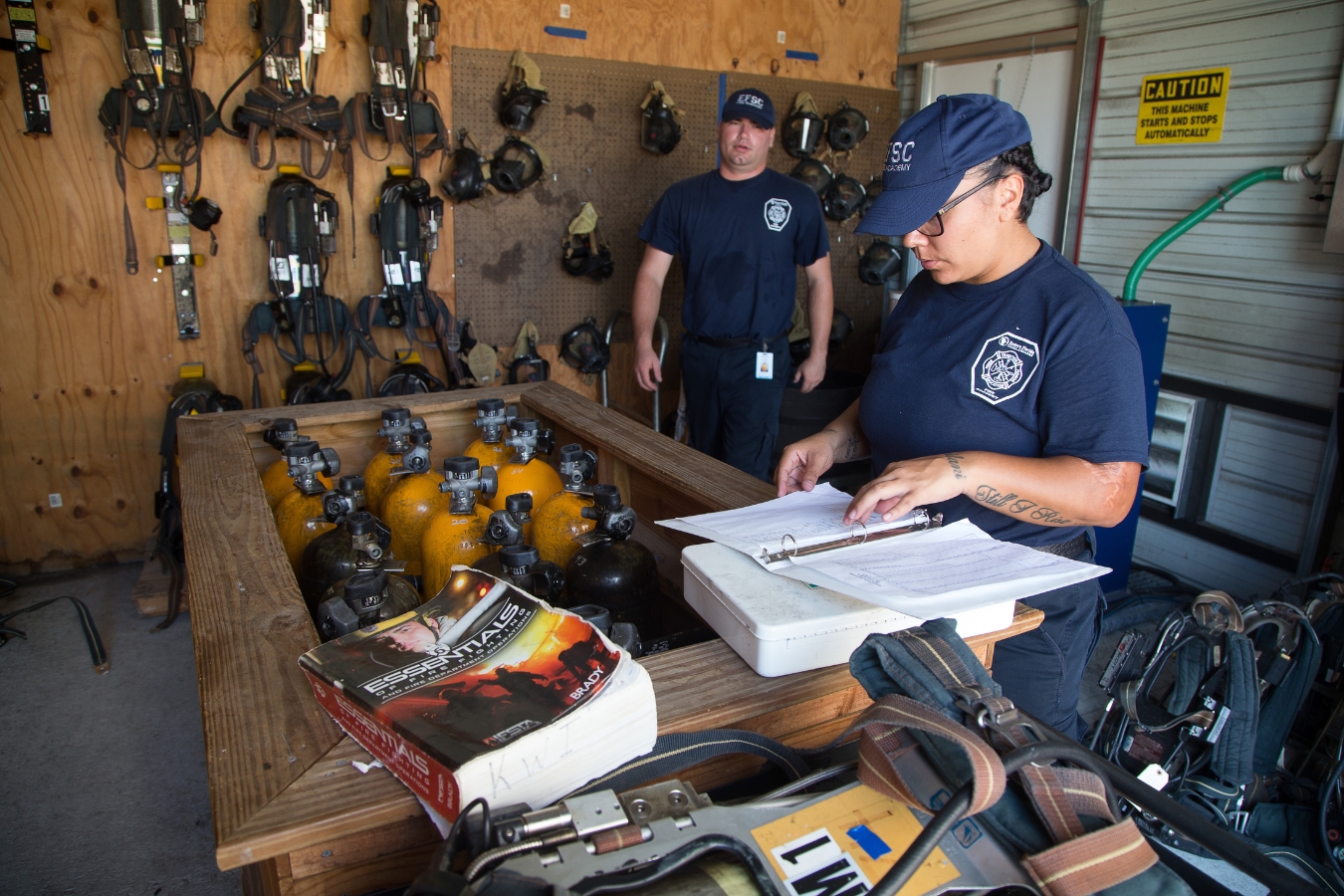 Two fire fighter students review their book materials as they work in a room with firefighting gear