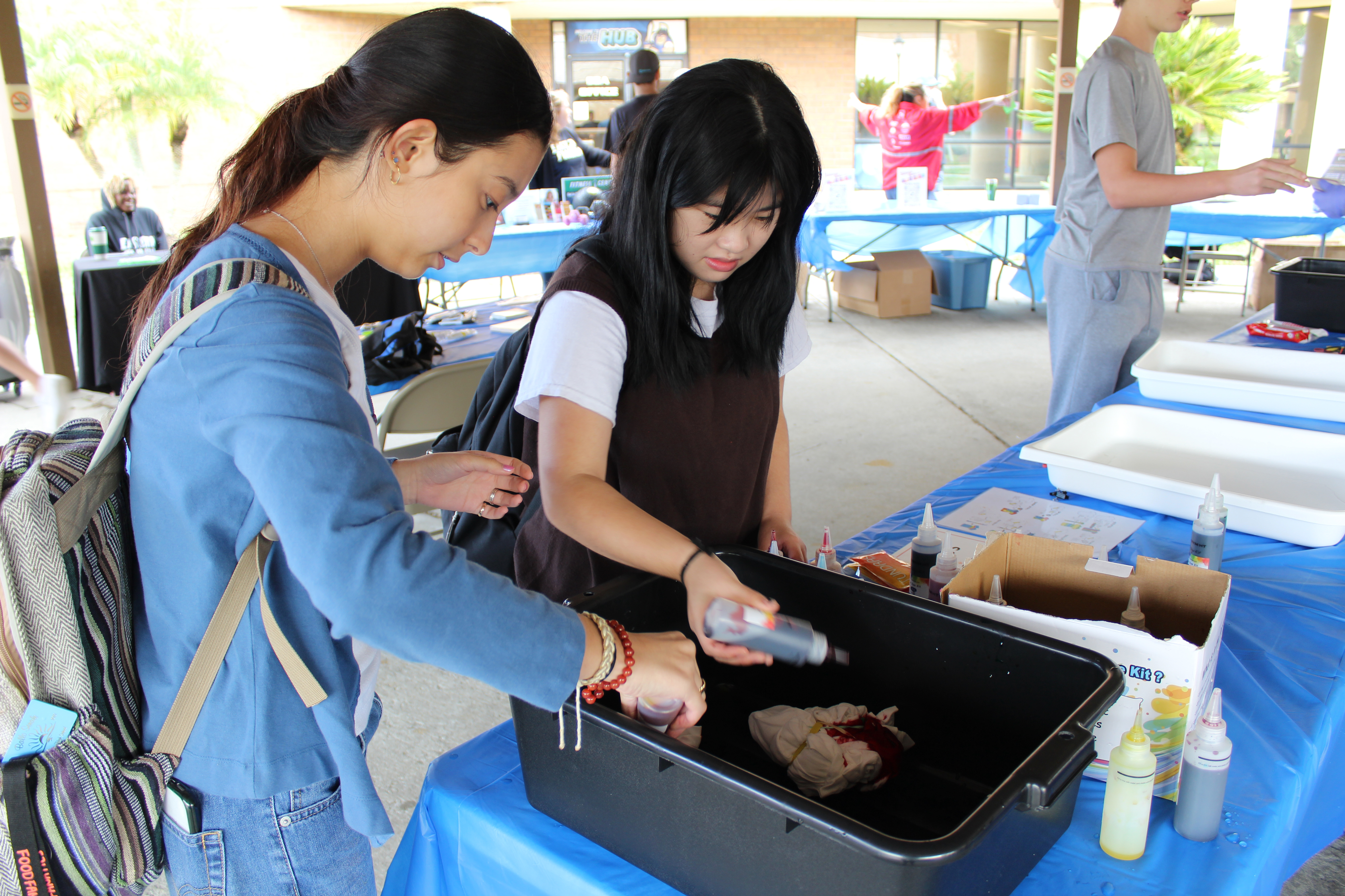 Two female students participating in a tie-dye activity at an outdoor student event, they are dying a tee shirt together.