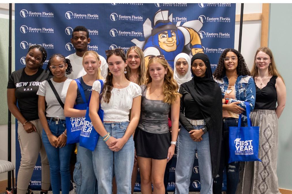 A group of diverse students stands in front of a Mr. Titan backdrop, holding First Year Experience tote bags.
