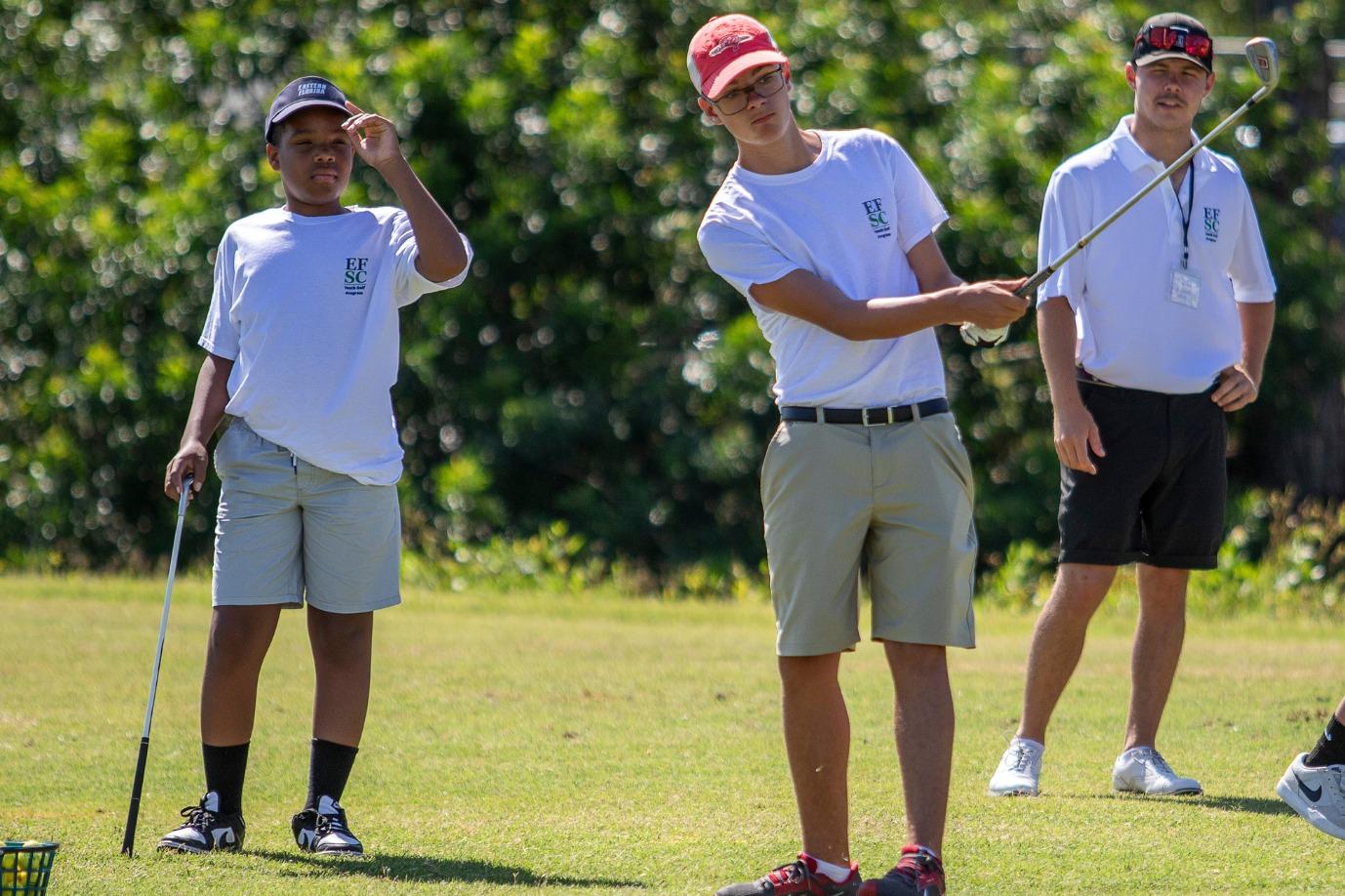 Young boy is swinging his golf club while a coach and another boy watch