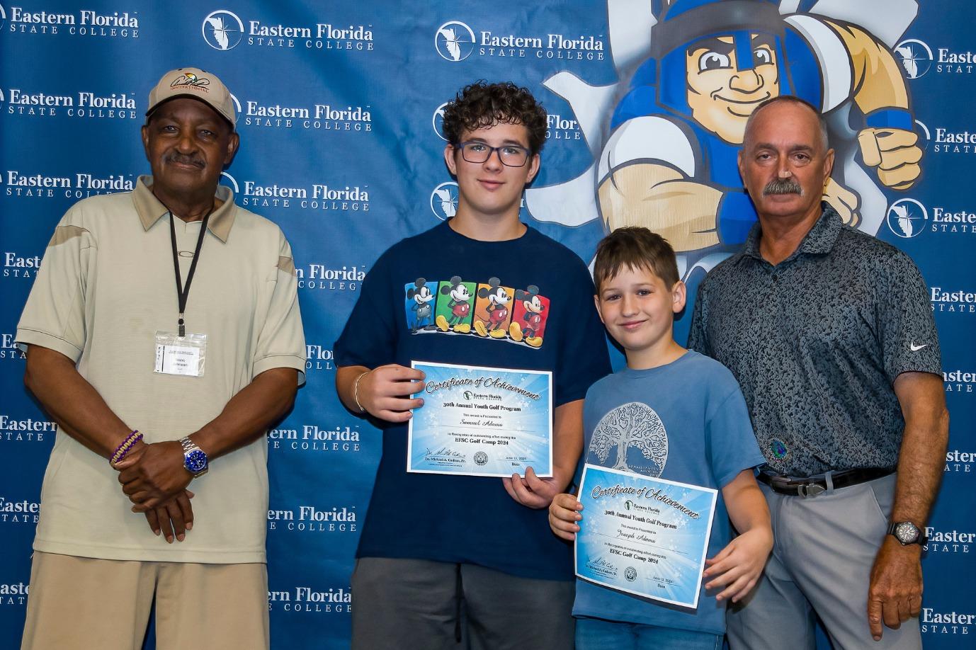Two participants are posing for a photo with the coaches during a celebratory event while holding certificates
