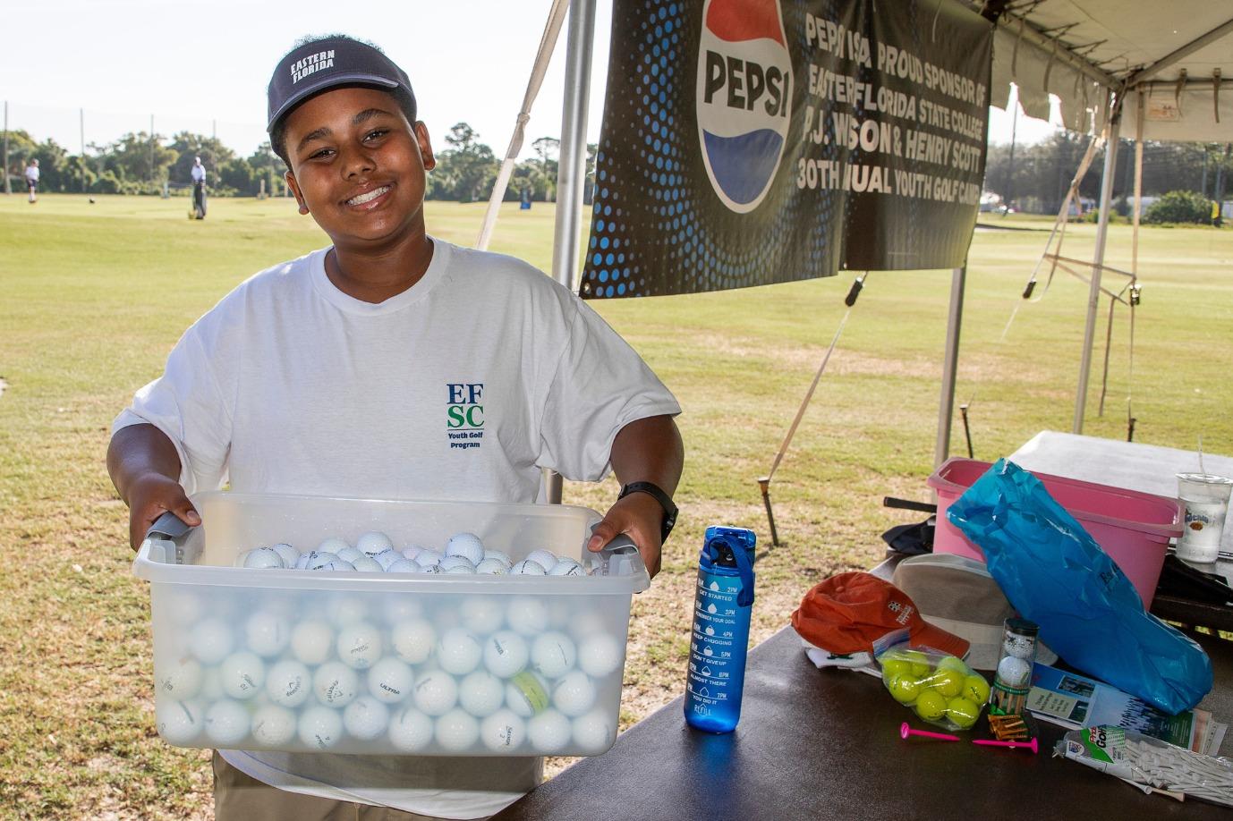 A young boy is smiling while holding a bucket of golf balls and wearing an EFSC tee shirt