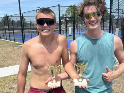 Two boys are smiling next to a volleyball court holding trophies