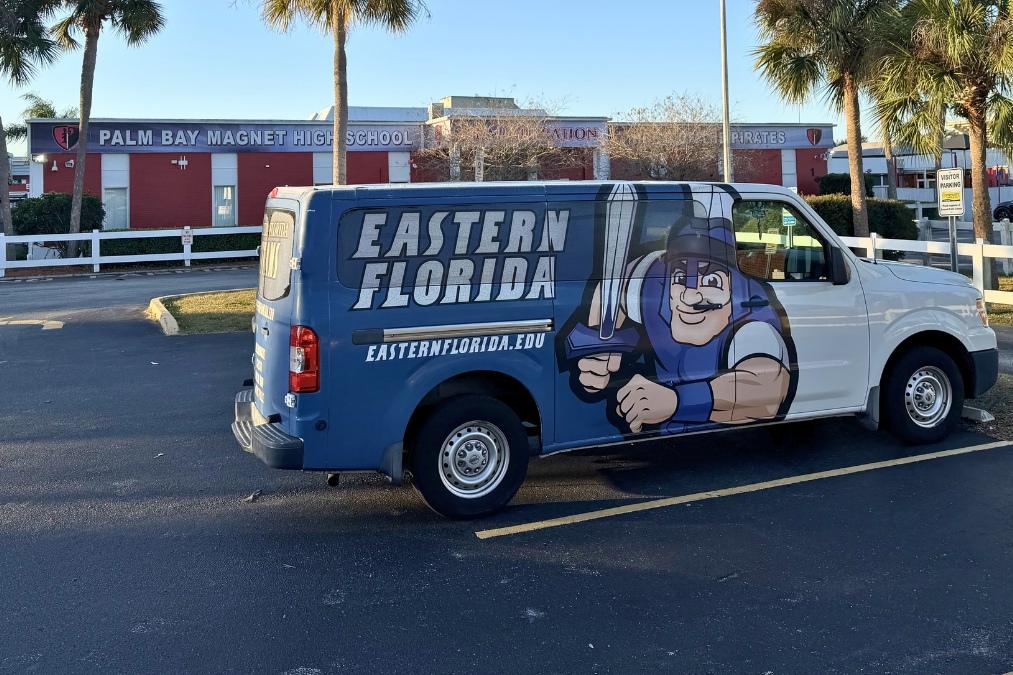 An Eastern Florida State College-branded van featuring a large knight graphic and the text “Eastern Florida” and “easternflorida.edu” is parked in a lot in front of Palm Bay Magnet High School, with the school building, palm trees, and signage visible in the background.