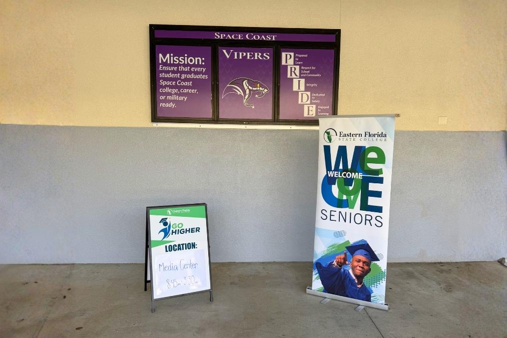 A “Go Higher” sign on a stand directing visitors to the Media Center (8:45–3:30) sits beside a vertical “Welcome Seniors” banner from Eastern Florida State College, displayed in a school hallway beneath a purple “Space Coast Vipers” mission and values wall sign.