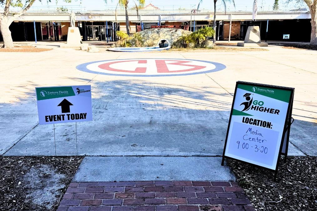 Two “Go Higher” directional signs placed along a walkway in a school courtyard—one with an arrow reading “Event Today,” and another on a stand listing the Media Center location (9:00–3:30)—with a large painted logo in the center of the courtyard, a decorative rock feature, and school buildings in the background.