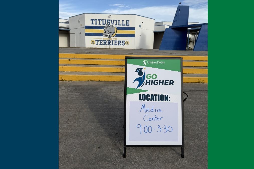 A “Go Higher” sign on a stand in front of Titusville High School, displaying directions to the Media Center (9:00–3:30), with the school building behind it featuring “Titusville Terriers” painted on the wall and yellow steps leading up to the entrance.