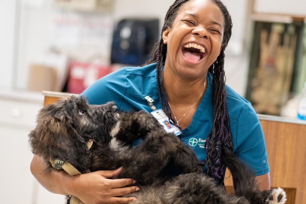 Student in teal Eastern Florida State College veterinary program scrubs laughs while holding a fluffy black dog in a classroom or clinic setting.