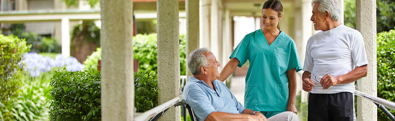 A woman in scrubs speaks with a man in a wheelchair while another man looks on.