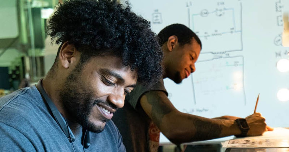Student works on an electronics or engineering project while another student writes notes beside a whiteboard covered with circuit diagrams.