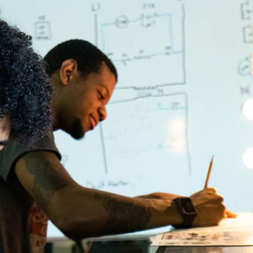 Student works on an electronics or engineering project while another student writes notes beside a whiteboard covered with circuit diagrams.