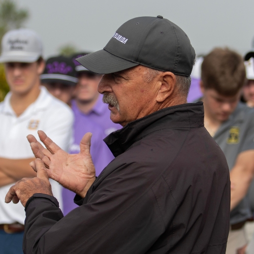 An image of Jamie Howell in a black jacket and dark baseball cap speaks to a group of golfers gathered outdoors, gesturing with both hands while participants in golf attire listen in the background on a golf course.