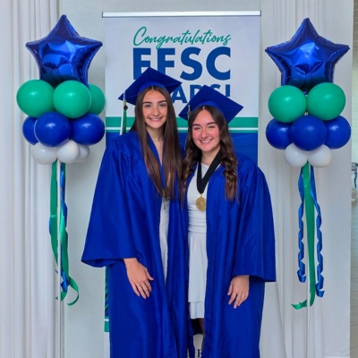 Madeleine and Gianna Dumonteil, young women with long brown hair wearing blue graduation gowns and gold medallions around their necks