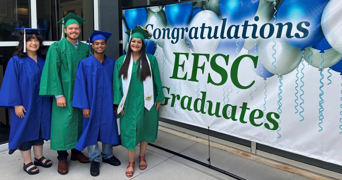 Four graduates wearing green and blue gowns stand by a large congratulations banner
