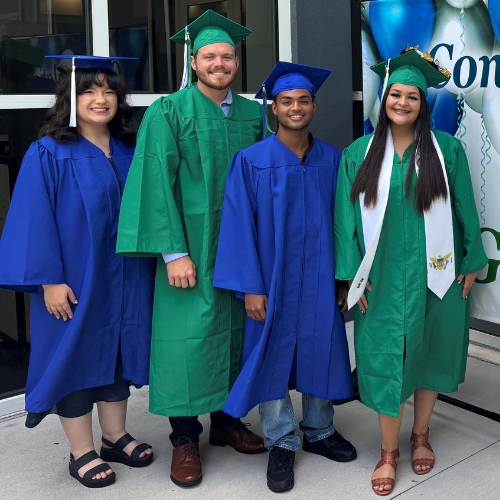 Four graduates in blue and green robes stand by a congratulatory banner
