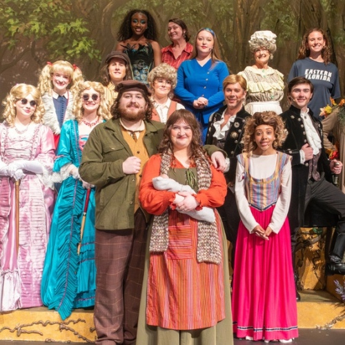 Large cast group poses onstage in costume for a theatrical production of “Into the Woods,” standing in front of a forest backdrop. The group wears a variety of colorful fairy tale-inspired outfits, including dresses, cloaks, hats, and formal jackets, while arranged in rows across the stage set.