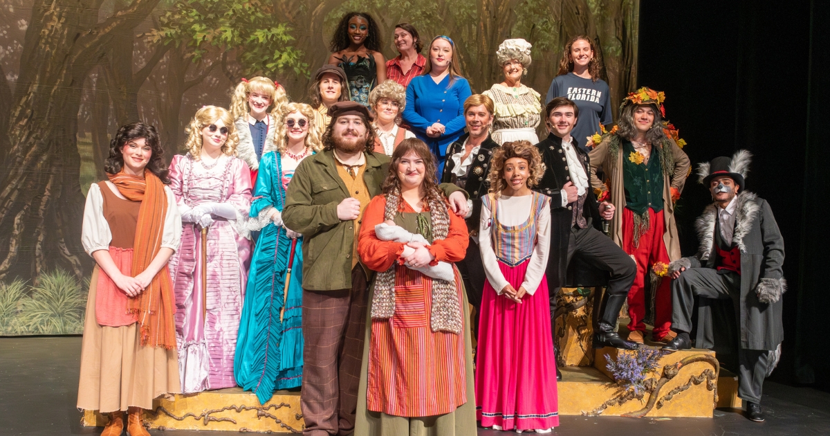 Large cast group poses onstage in costume for a theatrical production of “Into the Woods,” standing in front of a forest backdrop. The group wears a variety of colorful fairy tale-inspired outfits, including dresses, cloaks, hats, and formal jackets, while arranged in rows across the stage set.