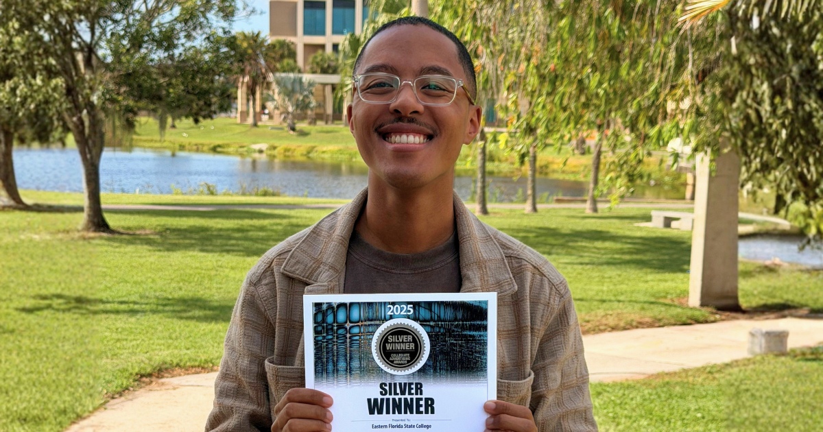 Josh Williams standing outdoors on a college campus holding a “Silver Winner” 2025 Collegiate Advertising Award certificate for Eastern Florida State College. A lake, palm trees, and campus buildings are visible in the background.