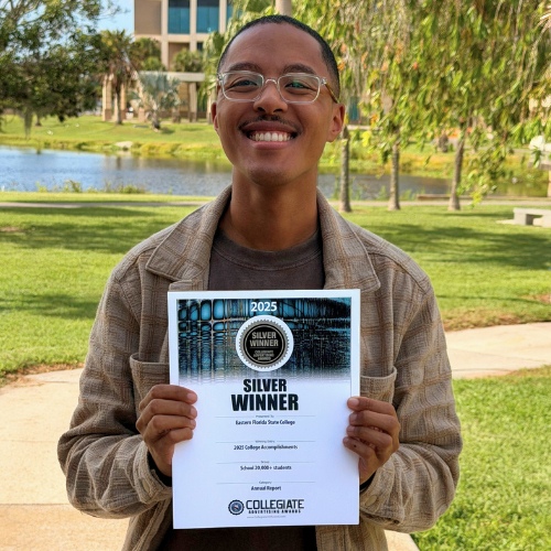 Josh Williams standing outdoors on a college campus holding a “Silver Winner” 2025 Collegiate Advertising Award certificate for Eastern Florida State College. A lake, palm trees, and campus buildings are visible in the background.