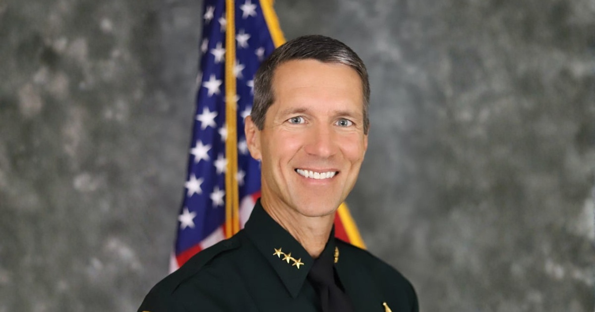 Formal portrait of Michael DeMorat in a dark green law enforcement uniform with gold star insignia and collar pins, standing in front of a blurred U.S. flag. He is facing forward and smiling, wearing a black tie and a nameplate on the chest, with a sheriff-style badge visible. The background is a neutral gray studio backdrop with soft lighting.