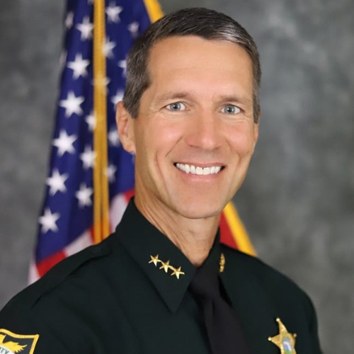 Formal portrait of Michael DeMorat in a dark green law enforcement uniform with gold star insignia and collar pins, standing in front of a blurred U.S. flag. He is facing forward and smiling, wearing a black tie and a nameplate on the chest, with a sheriff-style badge visible. The background is a neutral gray studio backdrop with soft lighting.